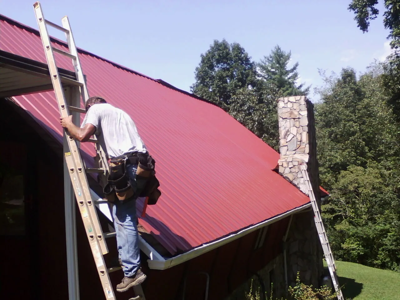 Roofer on a ladder beside a red metal roof and stone chimney — Soffit & Fascia Repair by Roofing Brothers in Batesville